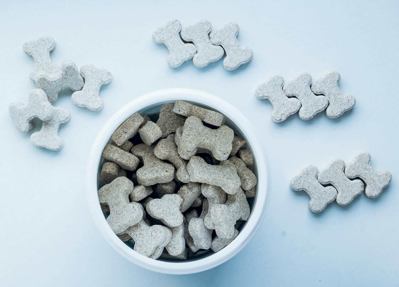 Top view of bone-shaped dog treats in a white bowl, on a light blue background.